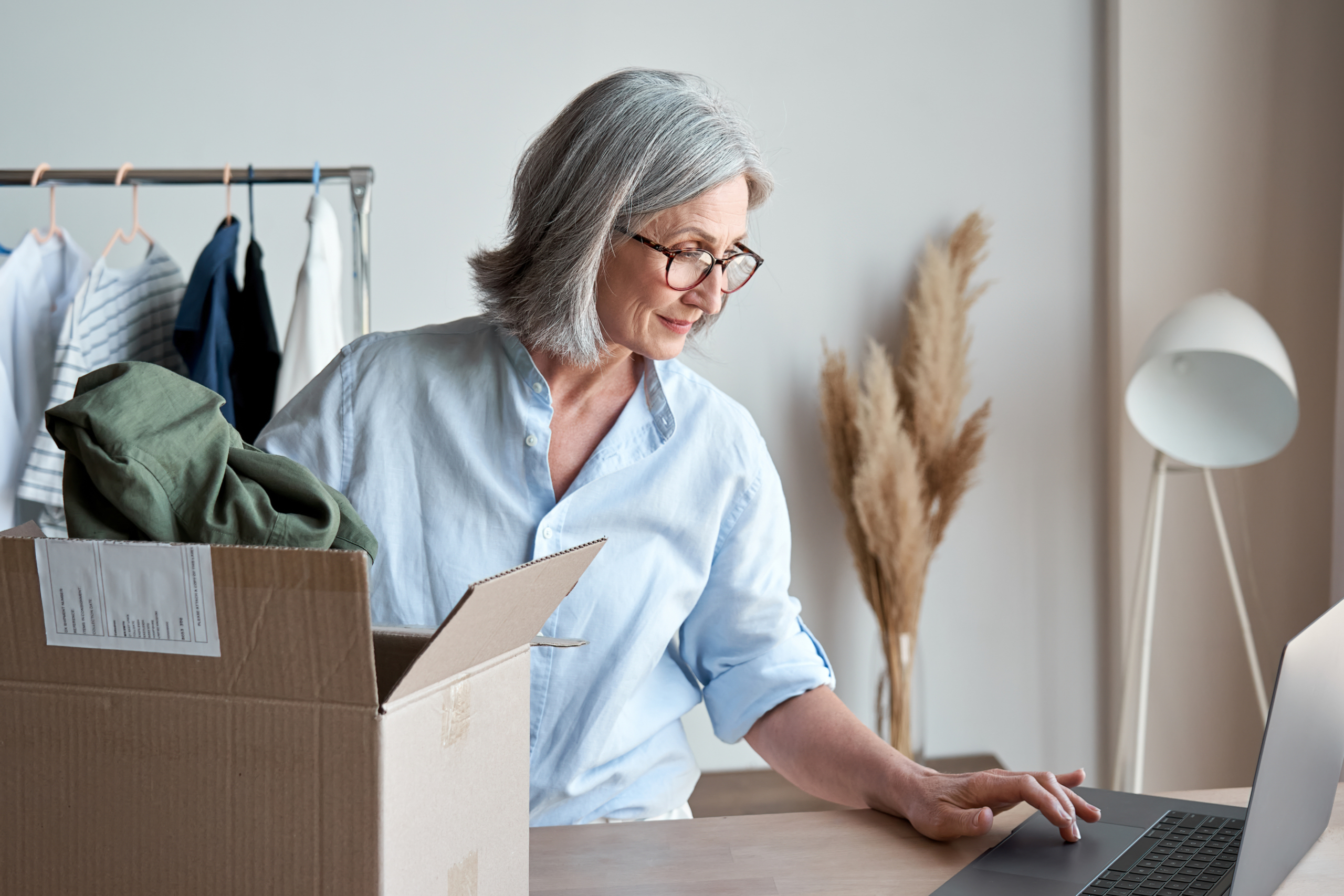 Mature woman fashion designer, dropshipping online clothing store small business owner looking for new e commerce order on laptop preparing packing in postal mail delivery shipping box at workplace. Mature woman fashion designer, dropshipping online clothing store small business owner looking for new e commerce order on laptop preparing packing in postal mail delivery shipping box at workplace.