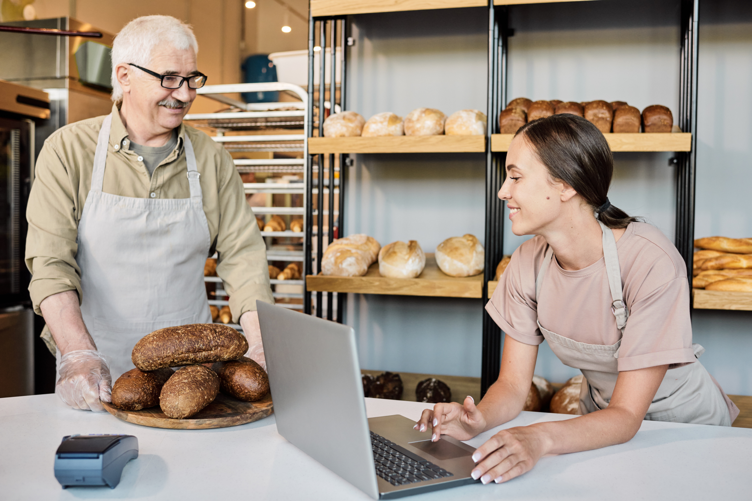 Happy woman with laptop looking at pile of fresh loaves of bread Happy woman with laptop looking at pile of fresh loaves of bread on wooden board held by mature baker